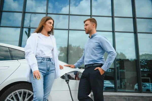 Happy young adult man and smiling woman charging electric car
