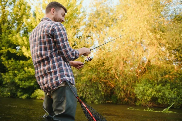 Man with fishing rod, fisherman men in river water outdoor. Catching trout fish in net. Summer fishing hobby.