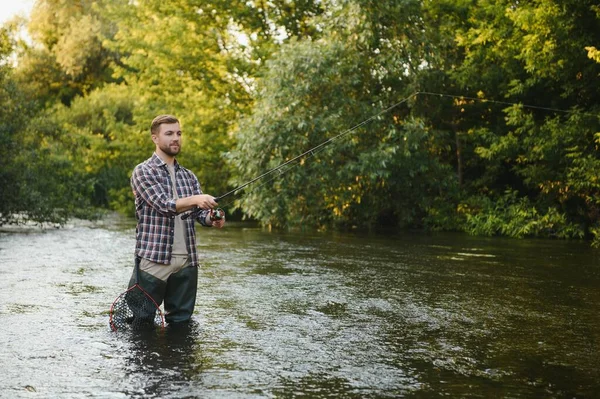 Man with fishing rod, fisherman men in river water outdoor. Catching trout fish in net. Summer fishing hobby.