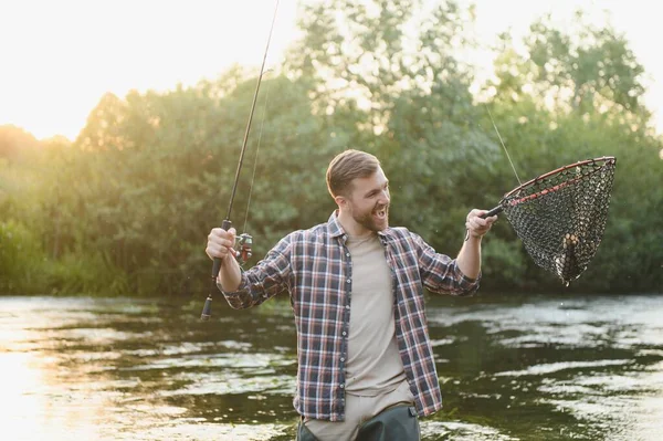 Man with fishing rod, fisherman men in river water outdoor. Catching trout fish in net. Summer fishing hobby.