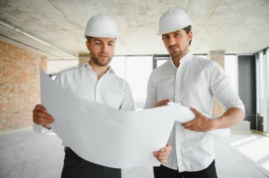 A front view of two smart architects with white helmets reviewing blueprints at a construction site on a bright sunny day.