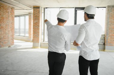 A front view of two smart architects with white helmets reviewing blueprints at a construction site on a bright sunny day.