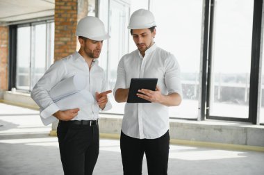 A front view of two smart architects with white helmets reviewing blueprints at a construction site on a bright sunny day.
