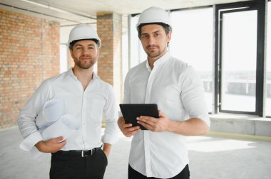 A front view of two smart architects with white helmets reviewing blueprints at a construction site on a bright sunny day.