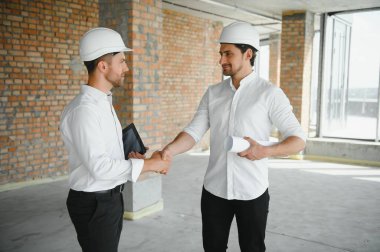 A front view of two smart architects with white helmets reviewing blueprints at a construction site on a bright sunny day.