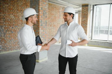 A front view of two smart architects with white helmets reviewing blueprints at a construction site on a bright sunny day.