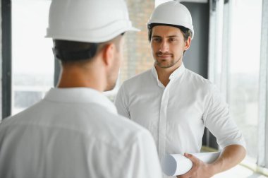 A front view of two smart architects with white helmets reviewing blueprints at a construction site on a bright sunny day.