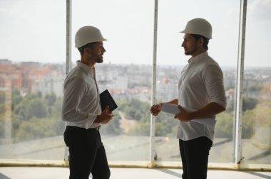 A front view of two smart architects with white helmets reviewing blueprints at a construction site on a bright sunny day.