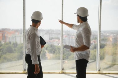A front view of two smart architects with white helmets reviewing blueprints at a construction site on a bright sunny day.