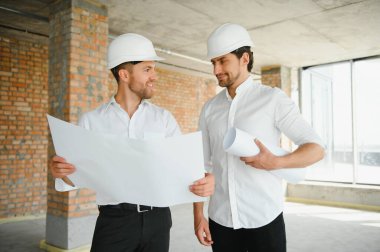 A front view of two smart architects with white helmets reviewing blueprints at a construction site on a bright sunny day.
