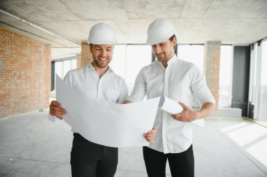 A front view of two smart architects with white helmets reviewing blueprints at a construction site on a bright sunny day.