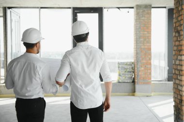 A front view of two smart architects with white helmets reviewing blueprints at a construction site on a bright sunny day.