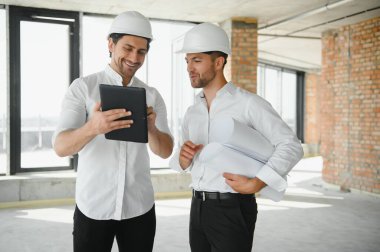 A front view of two smart architects with white helmets reviewing blueprints at a construction site on a bright sunny day.