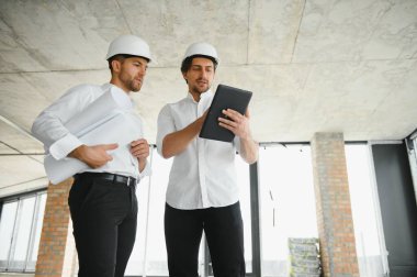 A front view of two smart architects with white helmets reviewing blueprints at a construction site on a bright sunny day.