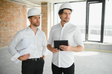 A front view of two smart architects with white helmets reviewing blueprints at a construction site on a bright sunny day.