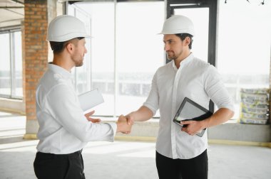 A front view of two smart architects with white helmets reviewing blueprints at a construction site on a bright sunny day.