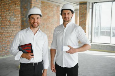 A front view of two smart architects with white helmets reviewing blueprints at a construction site on a bright sunny day.