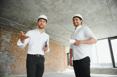 A front view of two smart architects with white helmets reviewing blueprints at a construction site on a bright sunny day.