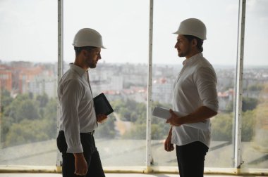 A front view of two smart architects with white helmets reviewing blueprints at a construction site on a bright sunny day.