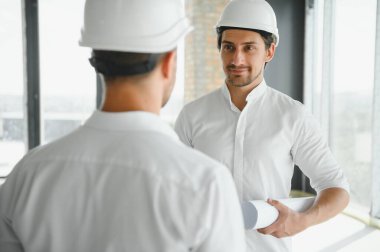 A front view of two smart architects with white helmets reviewing blueprints at a construction site on a bright sunny day.