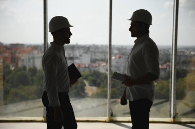 A front view of two smart architects with white helmets reviewing blueprints at a construction site on a bright sunny day.