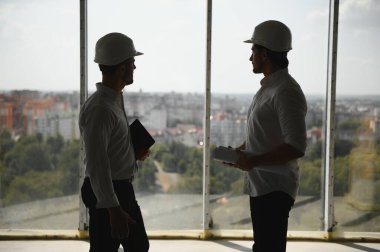 A front view of two smart architects with white helmets reviewing blueprints at a construction site on a bright sunny day.