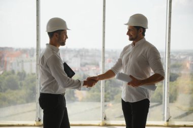A front view of two smart architects with white helmets reviewing blueprints at a construction site on a bright sunny day.