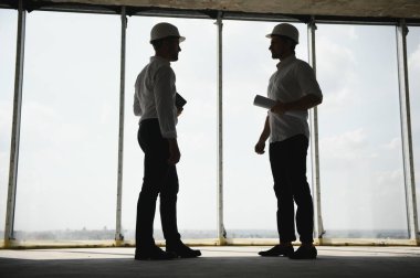 A front view of two smart architects with white helmets reviewing blueprints at a construction site on a bright sunny day.