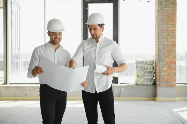 A front view of two smart architects with white helmets reviewing blueprints at a construction site on a bright sunny day.