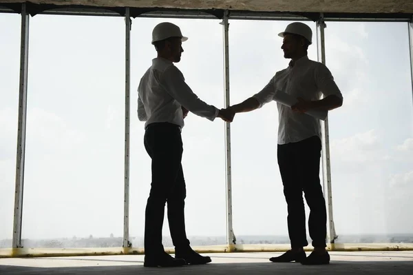 A front view of two smart architects with white helmets reviewing blueprints at a construction site on a bright sunny day.