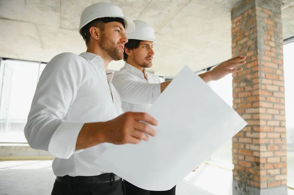 A front view of two smart architects with white helmets reviewing blueprints at a construction site on a bright sunny day.