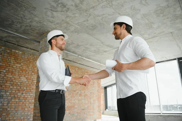 A front view of two smart architects with white helmets reviewing blueprints at a construction site on a bright sunny day.