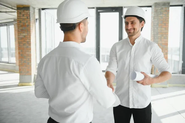 A front view of two smart architects with white helmets reviewing blueprints at a construction site on a bright sunny day.