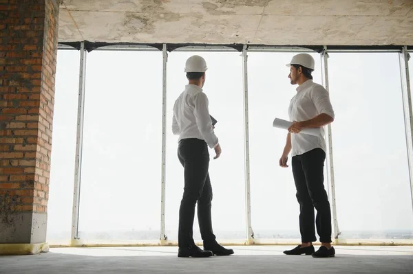 A front view of two smart architects with white helmets reviewing blueprints at a construction site on a bright sunny day.
