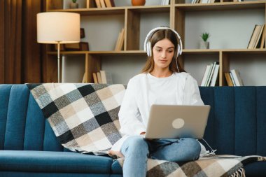 Beautiful young caucasian woman in casual clothing enjoying music and smiling while resting at home. Young woman with headphones using laptop at home.