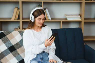 Portrait of attractive girl 20s wearing headphones using smartphone and listening to music while sitting on sofa at home.