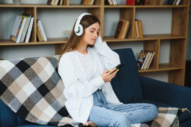Portrait of attractive girl 20s wearing headphones using smartphone and listening to music while sitting on sofa at home.