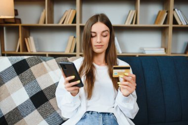 Mutlu genç evde kanepede oturarak online alışveriş yapıyor. Genç bir kadın akıllı telefonuyla internet sitesine kredi kartı numarasını giriyor.