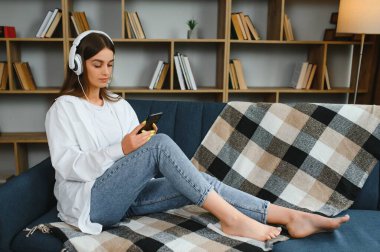 Portrait of attractive girl 20s wearing headphones using smartphone and listening to music while sitting on sofa at home.
