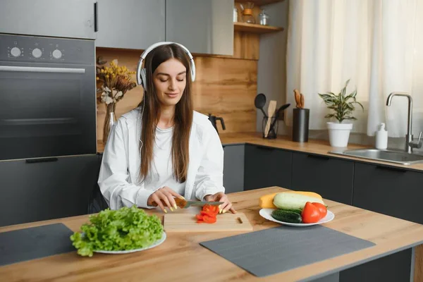 Funny young housewife woman in casual clothes preparing vegetable salad cooking food in light kitchen at home. Dieting healthy lifestyle concept. Listen music with headphones, sing song in cucumber.