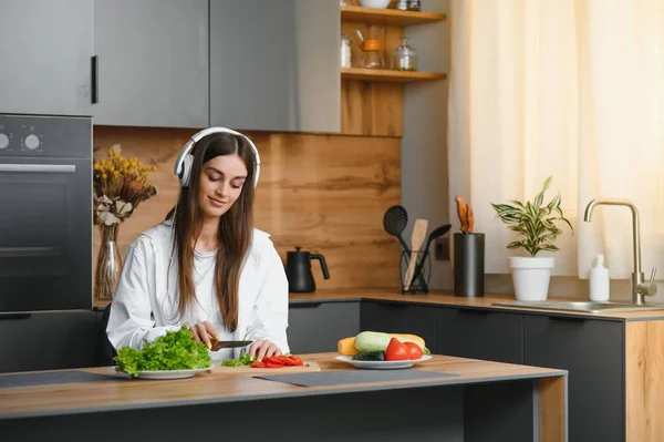 Funny young housewife woman in casual clothes preparing vegetable salad cooking food in light kitchen at home. Dieting healthy lifestyle concept. Listen music with headphones, sing song in cucumber.