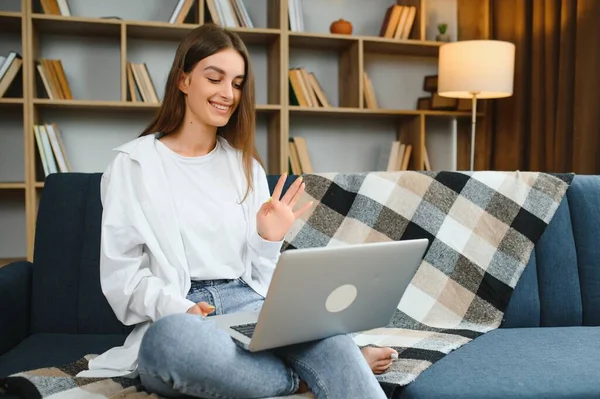 Smiling attractive young woman sitting on sofa using laptop communicating working online at home, happy teen girl typing on computer, enjoying writing blog or chatting with friends in social network.