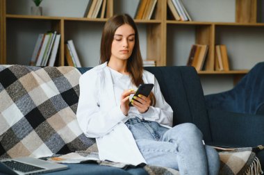 Young happy caucasian woman she 20s in casual clothes hold in hand use mobile cell phone sit on blue sofa indoor rest at home in own room apartment. People lifestyle concept.