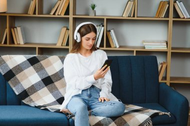 Portrait of attractive girl 20s wearing headphones using smartphone and listening to music while sitting on sofa at home.