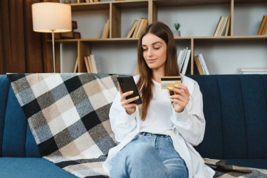 Elinde telefon ve kredi kartı olan mutlu genç bir kadın kanepede oturuyor. İnternetten alışveriş