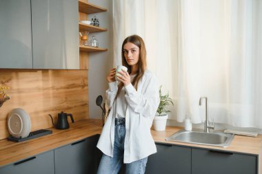 Happy young woman holding a cup of tea while standing in the kitchen at home in the morning. Healthy lifestyle concept