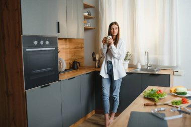 Happy young woman holding a cup of tea while standing in the kitchen at home in the morning. Healthy lifestyle concept