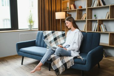 Dizüstü bilgisayar kullanan gülümseyen genç bir kadın, evdeki kanepede oturuyor, güzel bir kız alışveriş yapıyor ya da sosyal ağda sohbet ediyor, eğleniyor, film izliyor, bilgisayar projesinde serbest çalışıyor.