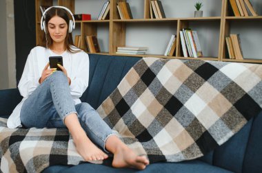 Beautiful young caucasian woman in casual clothing enjoying music and smiling while resting at home. Young woman with headphones using laptop at home.