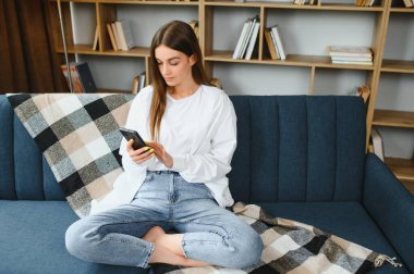 Young happy caucasian woman she 20s in casual clothes hold in hand use mobile cell phone sit on blue sofa indoor rest at home in own room apartment. People lifestyle concept.